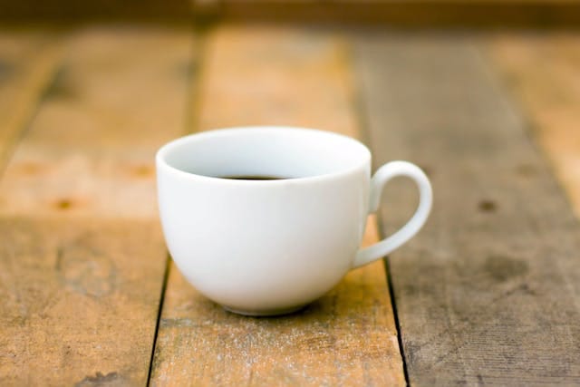 A single small white coffee cup, partially filled, on a rustic wood table.
