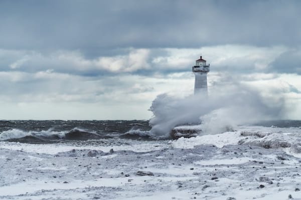 Rough waves crashing on a lighthouse on a snow-covered beach below thick gray clouds.